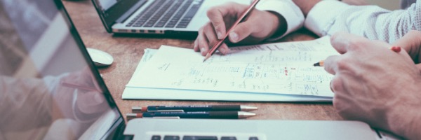 A cropped shot of two employees reviewing paper invoices, computer is in the foreground