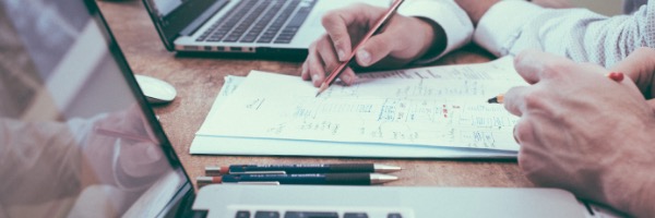 A cropped shot of two employees reviewing paper invoices, computer is in the foreground